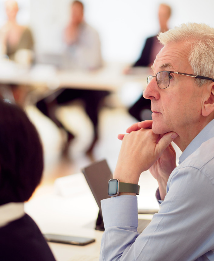 A close-up of an older man with glasses and a watch, watching a presentation